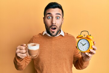 Young hispanic man holding coffee and alarm clock afraid and shocked with surprise and amazed...