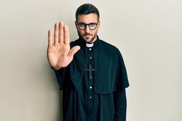 Young hispanic man wearing priest uniform standing over white background doing stop sing with palm of the hand. warning expression with negative and serious gesture on the face.