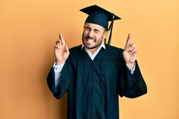 Young hispanic man wearing graduation cap and ceremony robe gesturing finger crossed smiling with hope and eyes closed. luck and superstitious concept.