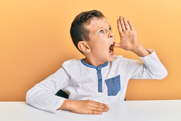 Adorable caucasian kid wearing casual clothes sitting on the table shouting and screaming loud to side with hand on mouth. communication concept.