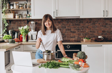 Close up woman in white t-shirt cooking soup with fresh vegetables in kitchen at home. Menu, recipe book banner. Girl reads the recipe in laptop. Caucasian model using internet in loft apartment.