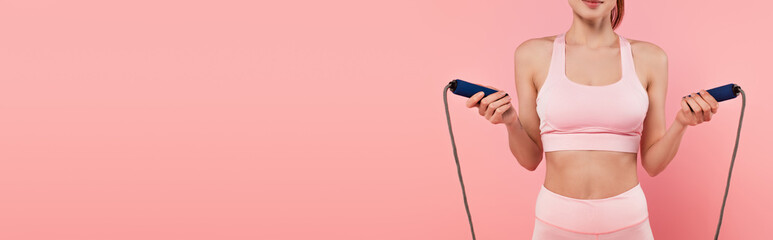 Cropped view of woman in sportswear holding jump rope isolated on pink, banner