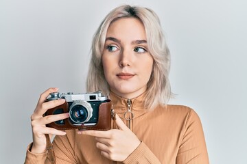 Young blonde girl holding vintage camera smiling looking to the side and staring away thinking.