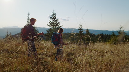 Woman and man trekking across grassy field. Backpackers using hiking poles