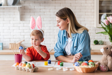 Happy mom and little son painting eggs for Easter in the kitchen