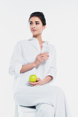 Young woman in white clothes holding fresh apple and glass of water isolated on white