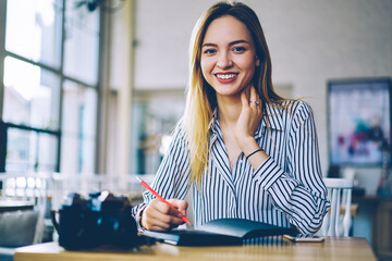 Obraz premium Portrait of cheerful Caucasian woman smiling at camera while spending leisure time for organisation planning in coffee shop, happy female student with education textbook posing at cafeteria table