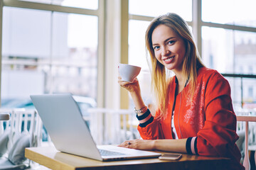 Portrait of cheerful female copywriter with cup of tasty coffee smiling at camera using modern...