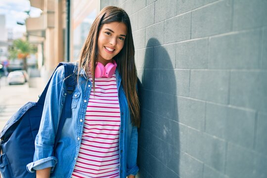 Young hispanic student girl smiling happy using headphones leaning on the wall at the city.