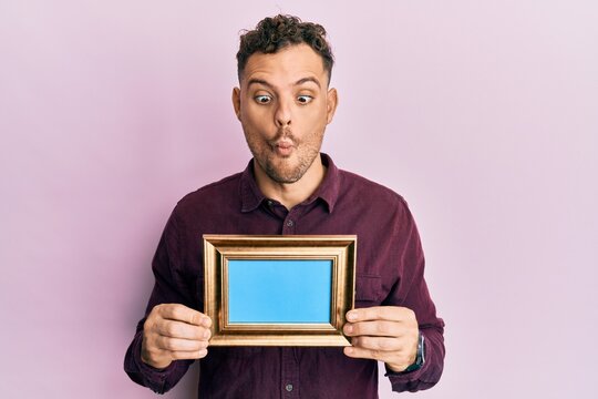 Young hispanic man holding empty frame making fish face with mouth and squinting eyes, crazy and comical.