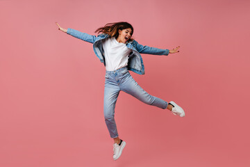 Trendy young woman jumping on pink background. Full length view of carefree female model in denim...