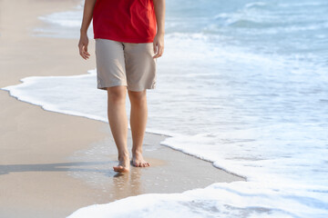 Young woman's bare feet with a wave coming and enjoying to travel on tropical beach in sunlight. Healthy female tan legs with droplets walking barefoot alone on sandy sea beach in summer. Copy space.