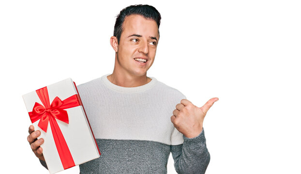 Handsome young man holding gift pointing thumb up to the side smiling happy with open mouth