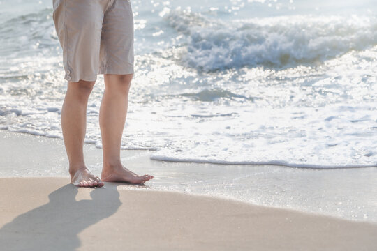 Young woman's bare feet with a wave coming and enjoying traveling to exotic nature. Beautiful healthy female legs with droplets standing barefoot alone on sandy sea beach in summer. Lifestyle concept