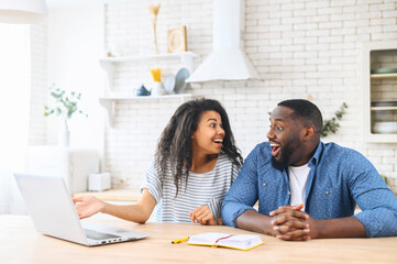 Excited multi ethnic couple in love does not believe their eyes, sits at the kitchen table in front to a laptop, looks to each other with shock, received mortgage approval by mail, win in lottery