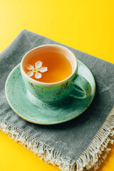 Turquoise cup of green or herbal tea on saucer with spring tree flowers on gray napkin over bright yellow background. Springtime concept. Vertical image.