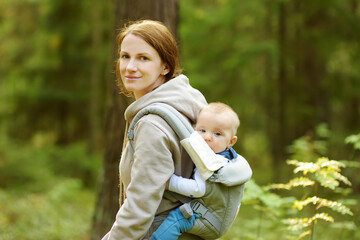 Young mother with her infant son in a baby carrier having fun hiking in a forest on beautiful summer day. New mom taking a walk with her baby son.