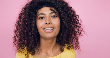happy young woman with curly hair looking at camera isolated on pink