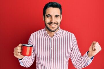 Young hispanic man holding coffee screaming proud, celebrating victory and success very excited with raised arm