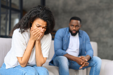 African-American young couple is arguing at home, a frustrated woman covered face with palms and crying, a sadness black man on the background. Newlywed have difficulties, relationship crisis