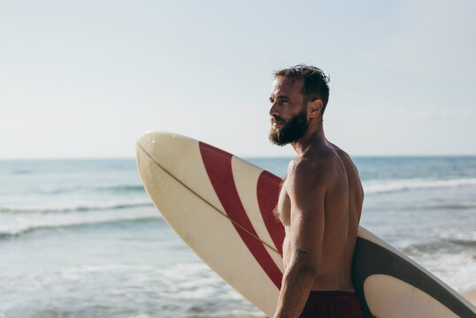Surfer holding his surfboard on the beach - Hipster man standing on the beach and waiting big waves for surfing - Fit bearded man training with surfboard to sea - Lifestyle and freedom concept