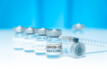 Covid-19 vaccine glass vials on the white surface with a blurred blue background and a syringe laying on top of one of the glass vials. 