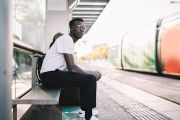 Serious dark skinned male in eyewear sitting on bus stop in shelter waiting for publica transport, handsome casually dressed hipster guy looking away for tramway spending time on urban settings - obrazy, fototapety, plakaty