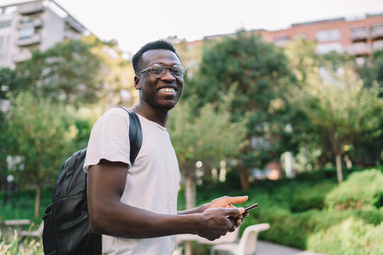 Cheerful black man smiling and looking at camera with smartphone on street