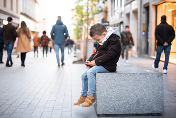 a little boy looking at the mobile in the city. a boy in a coat playing with the mobile