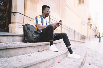 Smiling African American hipster guy in eyewear using cellular app for online phoning during travel vacations, happy male blogger in headphones checking received email and network publication