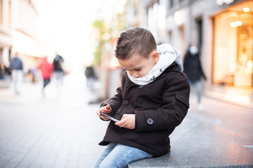 a little boy looking at the mobile in the city. a boy in a coat playing with the mobile