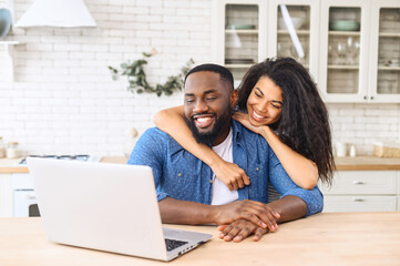 Cute multiracial couple enjoys leisure time at home with a laptop, happy woman and man in embrace look at the screen and laughs, watching videos, video calling with friends
