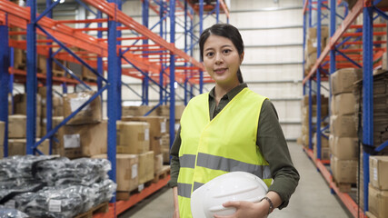 Portrait of smiling warehouse worker asian korean woman wearing safety vest and holding hard hat and looking in camera while standing in stockroom