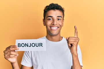Young handsome african american man holding bonjour french greeting word smiling with an idea or question pointing finger with happy face, number one © Krakenimages.com