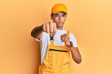 Young handsome african american man wearing handyman uniform over yellow background punching fist to fight, aggressive and angry attack, threat and violence