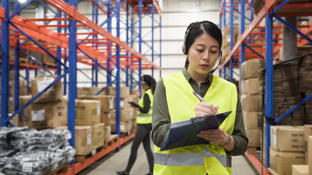 two young asian chinese girl customer service agent wearing headsets, talking online phone call and making note on clipboard in large distribution warehouse.