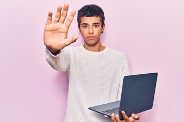 Young african amercian man holding laptop with open hand doing stop sign with serious and confident expression, defense gesture