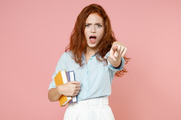 Young strict redhead student woman in blue shirt hold book point index finger camera on you command isolated on pastel pink background studio portrait Education high school university college concept.