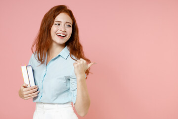 Young redhead student woman in blue shirt hold book point thumb finger aside on workspace mock up isolated on pastel pink background studio portrait Education high school university college concept