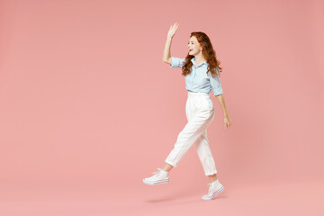 Full length body of young student friendly smiling happy redhead woman 20s in blue shirt pants walking going waving hand greeting loking back aside isolated on pastel pink background studio portrait.