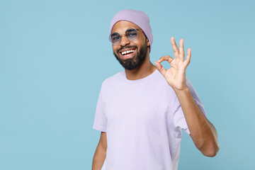 Young smiling happy positive satisfied hipster fun unshaven black dark-skinned african man 20s wear violet t-shirt hat glasses show ok okay gesture isolated on pastel blue background studio portrait.