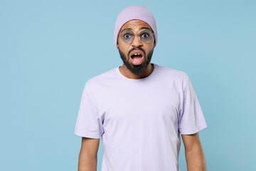 Young surprised shocked astonished impressed unshaven student black african man 20s in violet t-shirt hat glasses looking camera with opened mouth isolated on pastel blue background studio portrait.