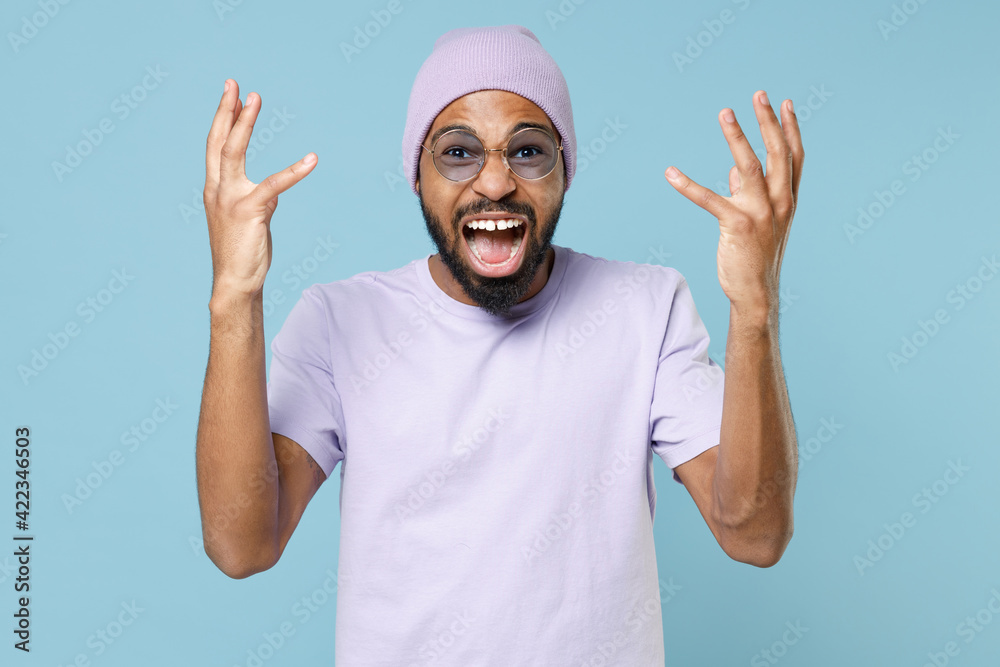 Wall mural Young irritated stressed nervous unshaven angry indignant black african man 20s wear violet t-shirt purple hat glasses spreading hands scream shout isolated on pastel blue background studio portrait.