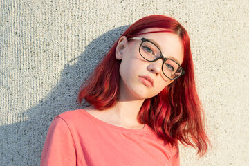 A teenage girl with dyed red hair, glasses and freckles. City portrait at sunset.