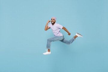 Fototapeta na wymiar Full length of young smiling positive confident black african man 20s in violet t-shirt hat glasses jump high run fast do winner gesture clench fist isolated on pastel blue background studio portrait.
