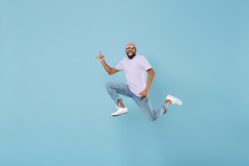 Obraz premium Full length young overjoyed black african man in violet t-shirt hat glasses jump high do horns up gesture depict heavy metal rock sign rock-n-roll isolated on pastel blue background studio portrait.