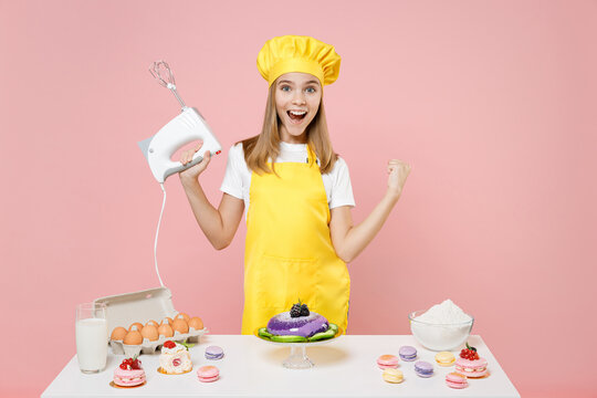 Teen girl chef cook confectioner baker in yellow apron toque cap at table hold kitchen mixer do winner gesture clench fist isolated on pink background studio Mousse cake food process workshop concept.