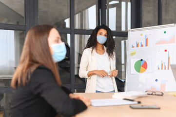 Multiethnic work group brainstorming in the contemporary office during pandemic period, a diverse colleagues wearing medical mask indoor. An African-American woman with covered face near whiteboard