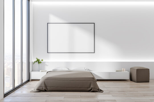 Blank white poster in black picture frame on white wall in stylish bedroom with huge window with city view, bed on wooden floor and brown pouf. Mock up