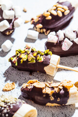 Bananas in dark chocolate with different toppings on stick, gray background. Healthy vegan dessert concept.